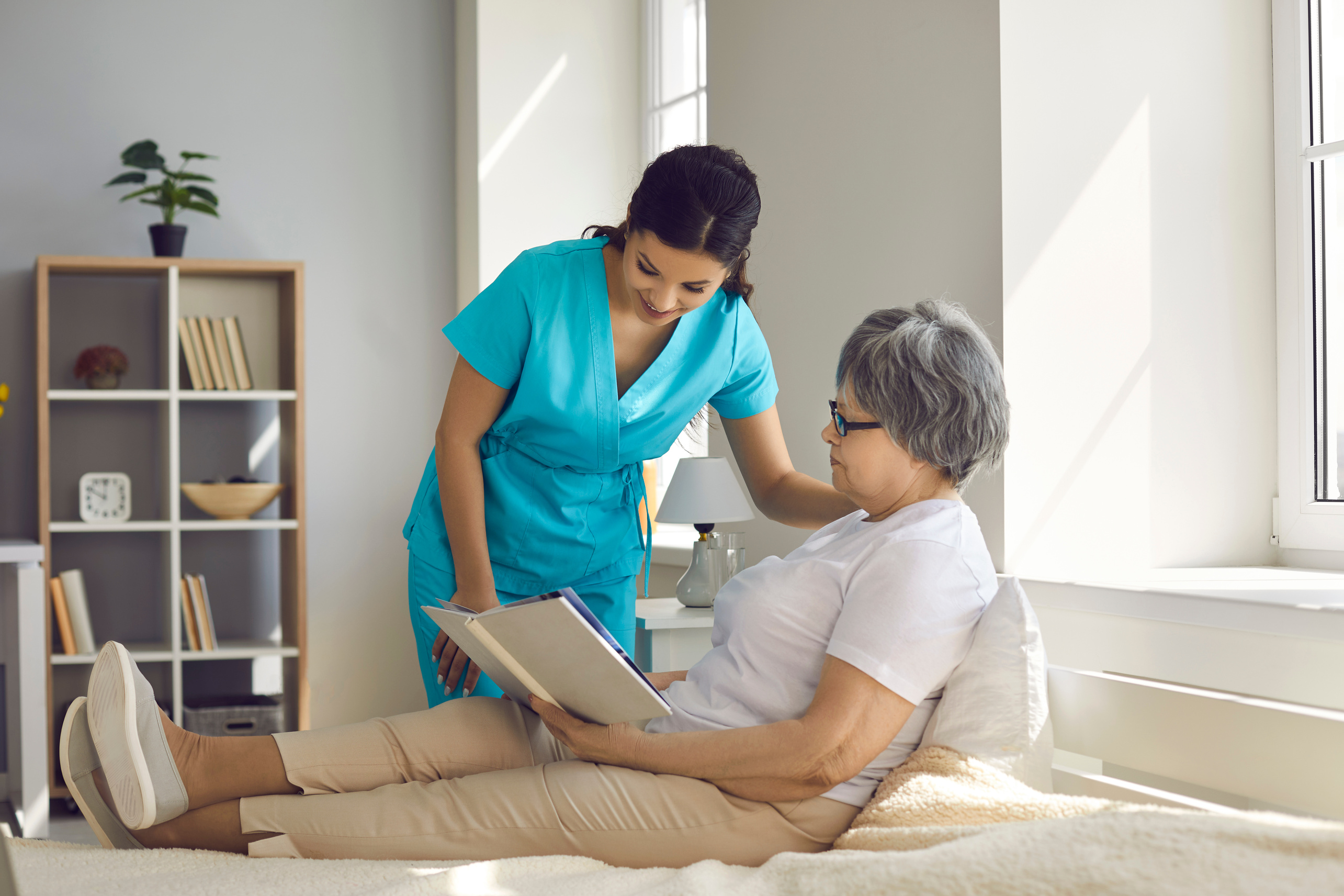 Home Care Nurse Talking to Relaxed Senior Woman Who's Sitting on Bed and Reading Book